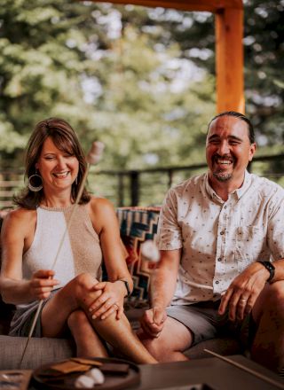 A couple smiling and sitting on an outdoor sofa, under a wooden pergola with greenery in the background.