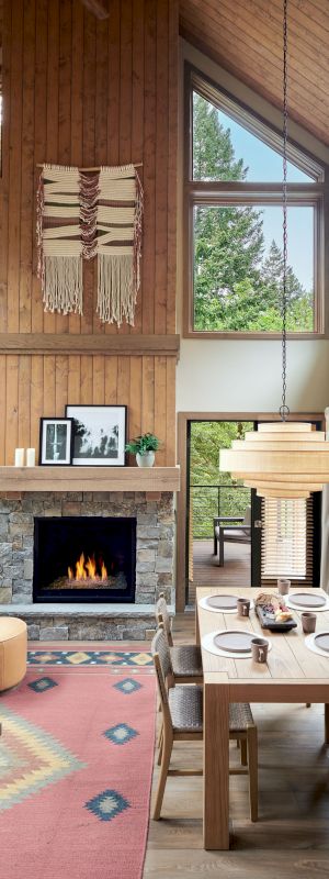 A cozy living room with a fireplace, dining table, large windows, and natural wood accents, creating a warm and inviting atmosphere.
