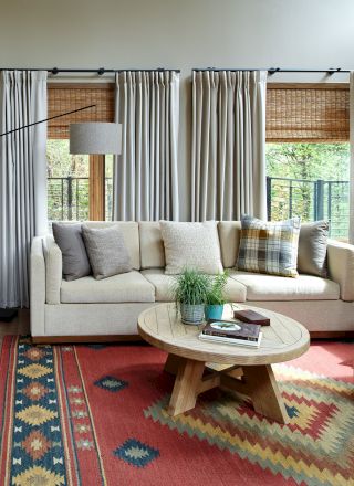 A cozy living room with a white sofa, plaid pillows, wooden coffee table, woven baskets, and a red patterned rug. Sunlight streams in through windows.