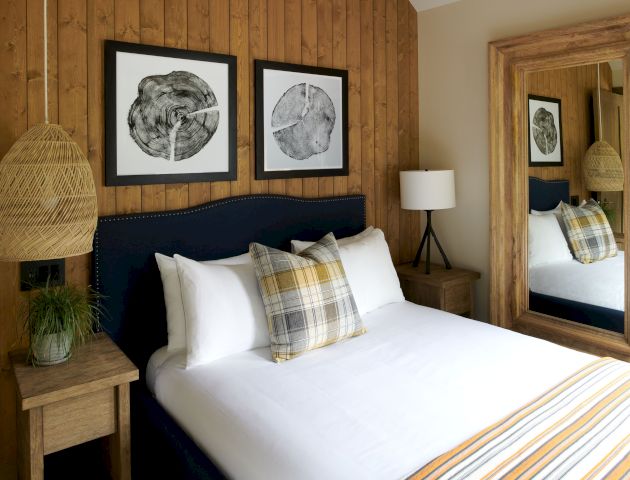 A cozy bedroom with a wooden wall, framed prints, a bed with plaid pillows, striped blanket, wicker lamp, side tables, and a large mirror.