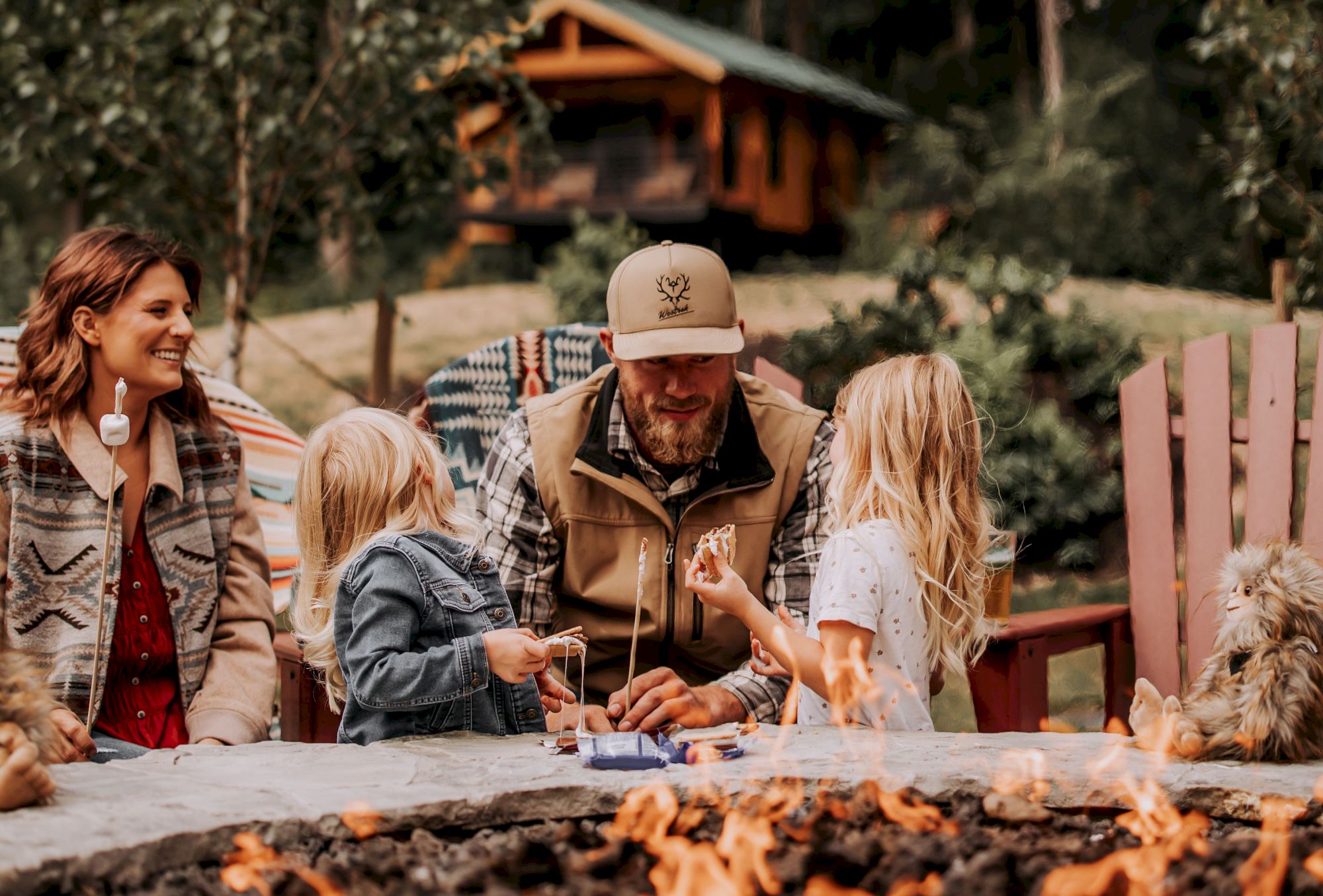 A family sits around a fire pit, enjoying a cozy outdoor setting with trees and a cabin in the background.