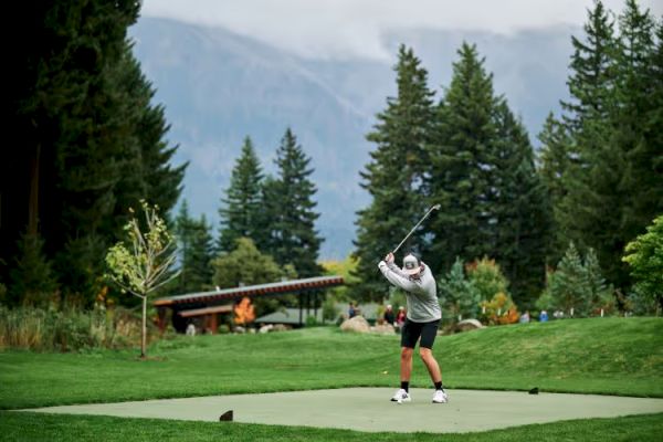 A golfer is mid-swing on a lush green course, surrounded by trees and mountains in the background.