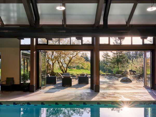 The image shows an indoor pool area with large windows overlooking an outdoor patio with seating and a beautiful garden view.