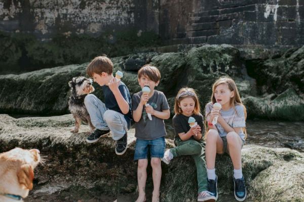 Four kids sit outdoors enjoying ice cream, accompanied by two dogs on a grassy area.