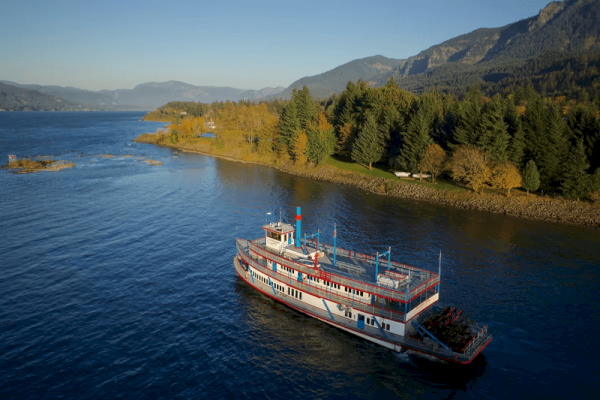 A paddleboat is cruising along a scenic river with lush forests and mountains in the background, under a clear blue sky.