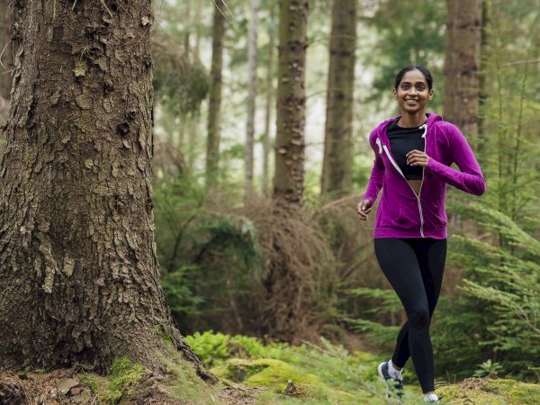 A person jogging in a forest, wearing a purple jacket and black leggings, surrounded by lush green trees and plants.