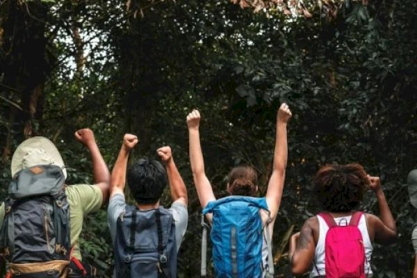 A group of hikers with backpacks face a lush forest, raising their arms in celebration or excitement, surrounded by dense greenery and trees.