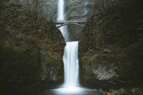 A waterfall flows with a bridge crossing above it, surrounded by lush vegetation and rocky cliffs.