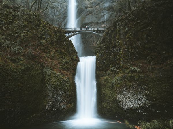 A waterfall flows with a bridge crossing above it, surrounded by lush vegetation and rocky cliffs.