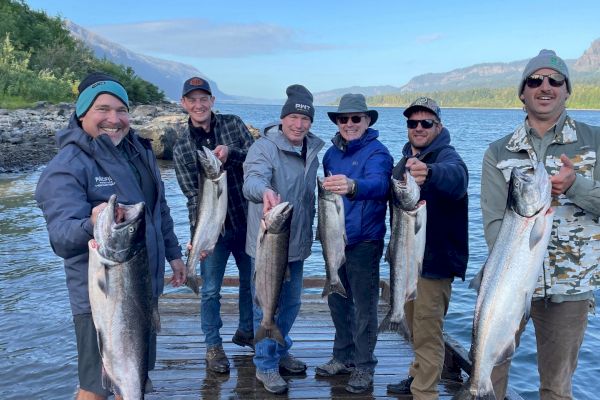 A group of six people standing on a dock by a river, each holding a large fish, smiling and dressed for outdoor weather.