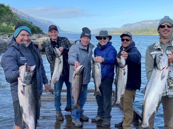 A group of six people standing on a dock by a river, each holding a large fish, smiling and dressed for outdoor weather.