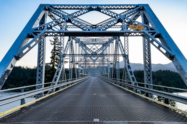A steel truss bridge spans a river, with trees and mountains in the background under a clear sky.