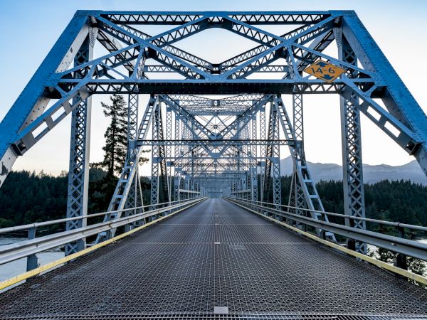 A steel truss bridge spans a river, with trees and mountains in the background under a clear sky.