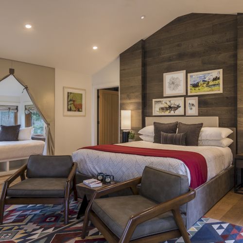 A cozy bedroom features a large bed, armchairs, rustic decor, and a window seat with cushions, all in a warm, inviting ambiance.