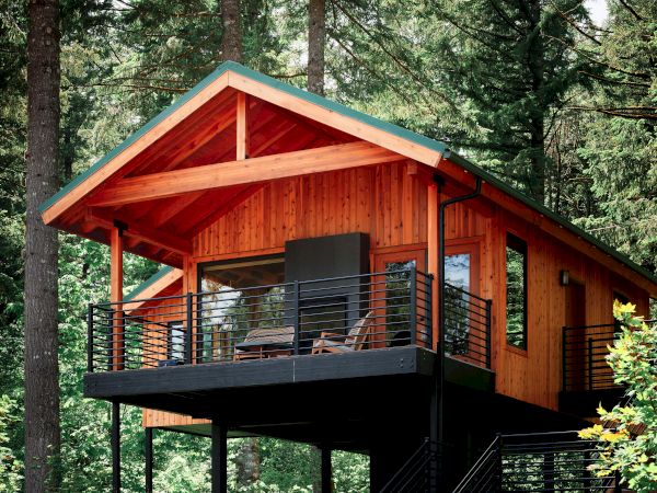 A modern elevated wood cabin with a deck and staircase, surrounded by tall trees in a forest setting.