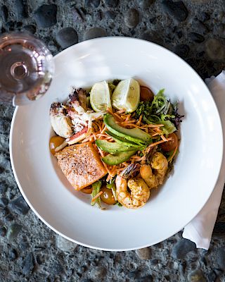 A seafood salad with salmon, shrimp, avocado, and lime wedges in a white bowl, paired with a glass of rosé wine.