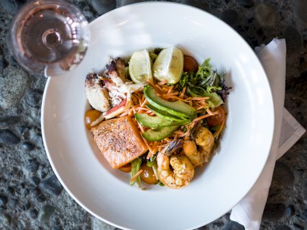 A seafood salad with salmon, shrimp, avocado, and lime wedges in a white bowl, paired with a glass of ros&eacute; wine.