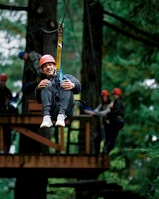 A person with a helmet is zip-lining through a forest while others watch from a wooden platform.
