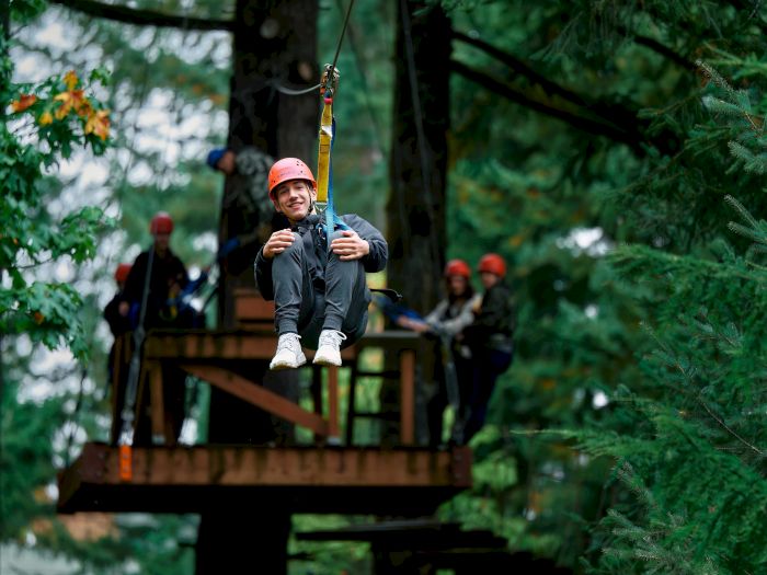 A person with a helmet is zip-lining through a forest while others watch from a wooden platform.