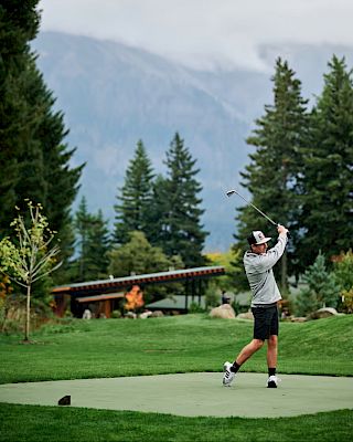 A person is playing golf on a lush green course set against a backdrop of mountains and trees, swinging a club.