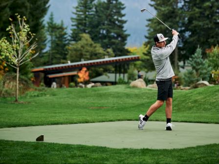 A person is playing golf on a lush green course set against a backdrop of mountains and trees, swinging a club.