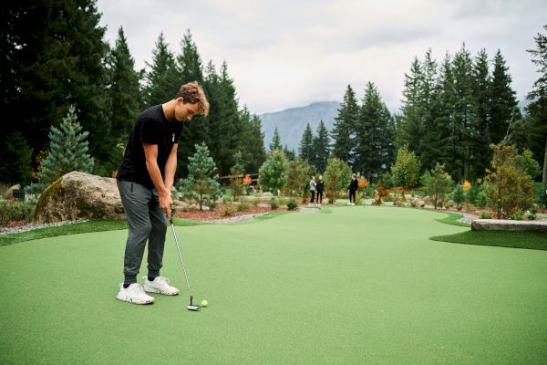 A person is playing mini-golf on a green course surrounded by trees, with mountains in the background under an overcast sky.