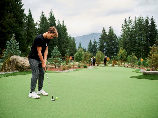 A person is playing mini-golf on a green course surrounded by trees, with mountains in the background under an overcast sky.
