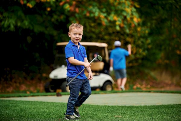 A young boy holding a golf club stands on grass, with a man and golf cart in the background amidst green foliage.