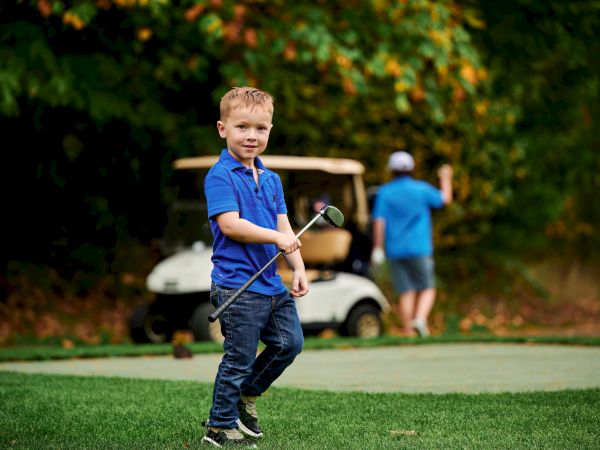 A young boy holding a golf club stands on grass, with a man and golf cart in the background amidst green foliage.