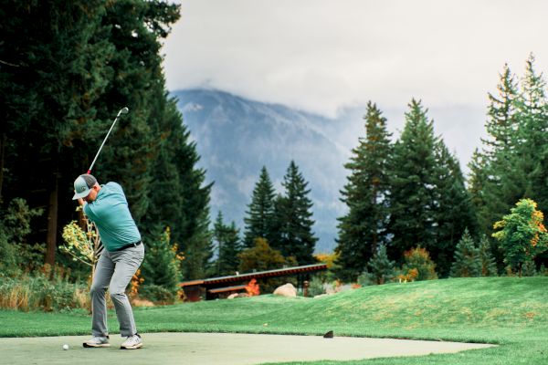 A person is swinging a golf club on a course surrounded by trees and mountains under a cloudy sky.