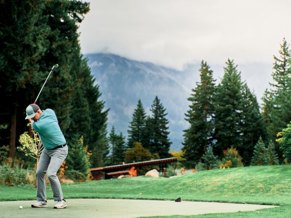 A person is swinging a golf club on a course surrounded by trees and mountains under a cloudy sky.
