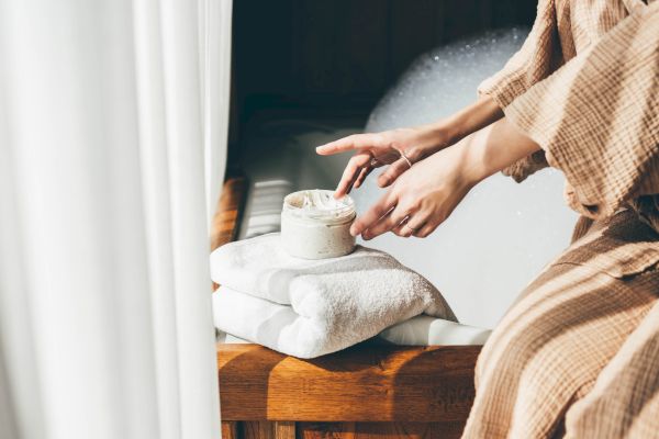 A person in a robe applies cream from a jar on a towel by a bubble-filled bathtub, creating a relaxing spa-like atmosphere.