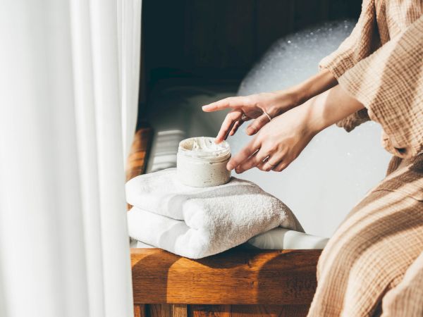 A person in a robe applies cream from a jar on a towel by a bubble-filled bathtub, creating a relaxing spa-like atmosphere.