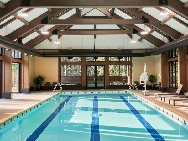 An indoor swimming pool with blue lane markings, wooden beams, skylights, and lounge chairs by the side in a spacious room.