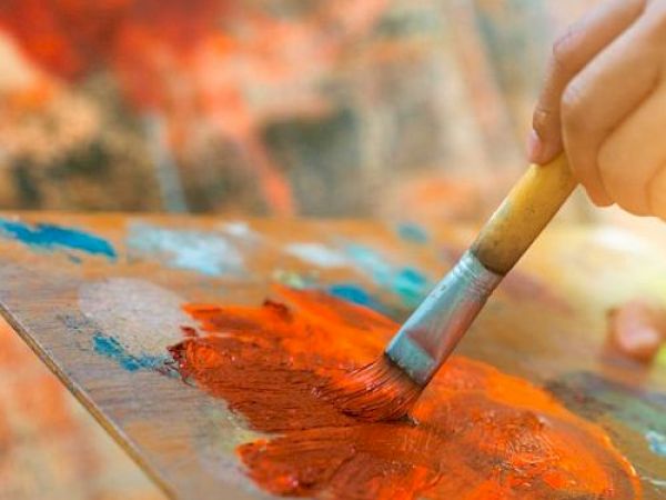 A hand is using a brush to mix vibrant red and orange paints on a palette, with a blurred, colorful background.