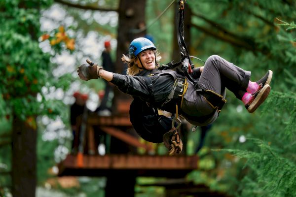 A person enjoying a zipline ride through a forest, wearing safety gear and making a thumbs-up gesture while others wait in the background.