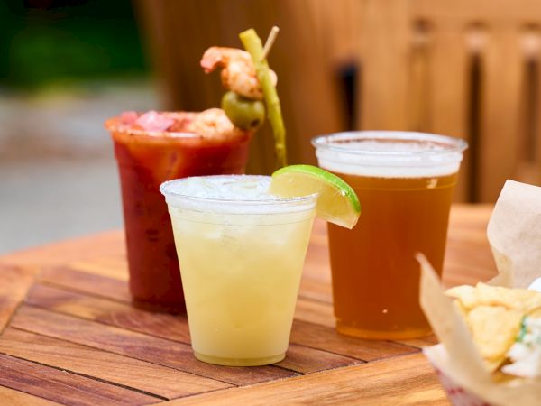 Three drinks are on a wooden table: a Bloody Mary with garnishes, a lime-garnished cocktail, and a glass of beer in plastic cups.