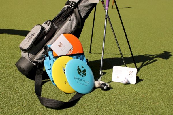 A golf bag, assorted discs, golf clubs, and a scorecard are on a green surface in the image.
