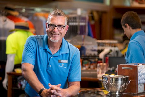 A smiling person in a blue polo stands in a shop with another person, surrounded by clothing and merchandise.