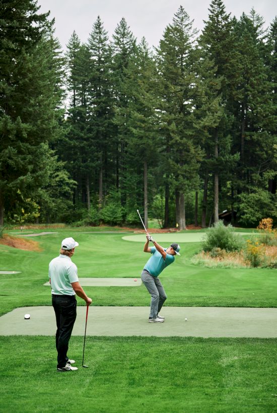 Two golfers on a green tee box, surrounded by trees, with one preparing to swing a club while the other watches.