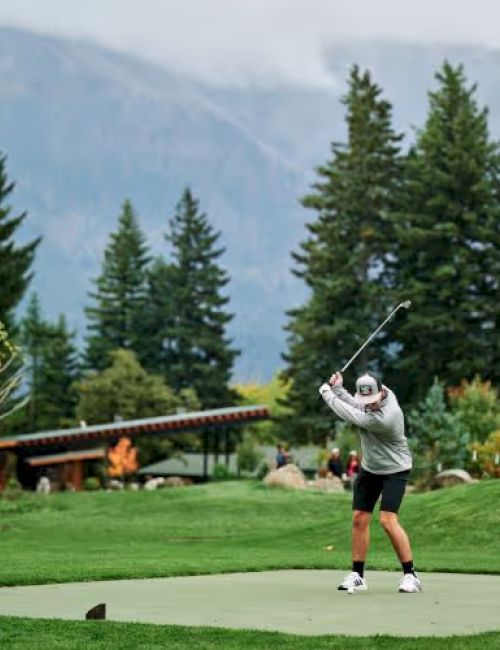 A person is playing golf on a green course surrounded by tall trees and mountains in the background.