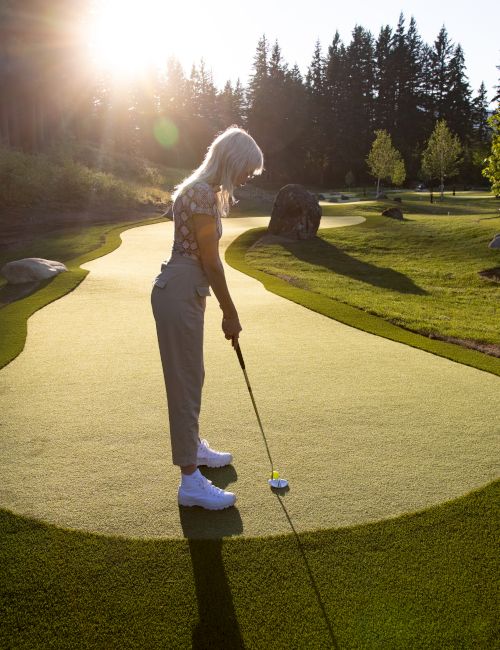 A person is playing golf on a sunny course surrounded by trees and grass, with the sun setting in the background.