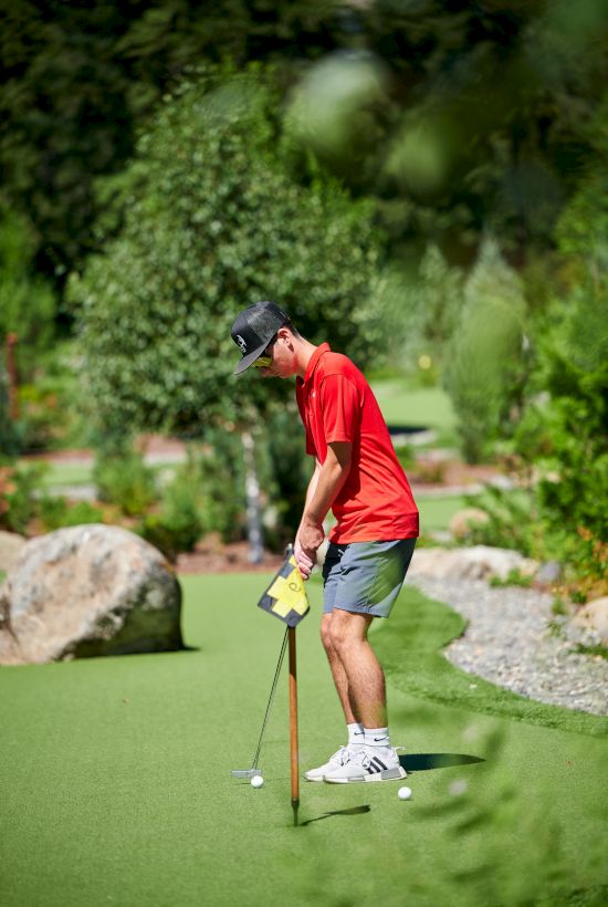 A person wearing a red shirt, shorts, and a cap is playing mini-golf on a green course with trees and rocks around.