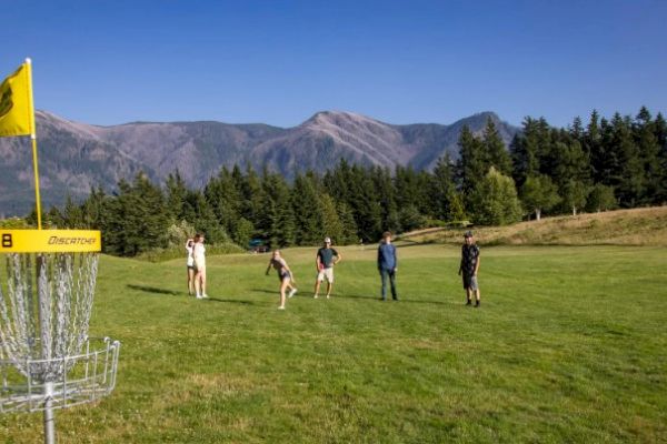 A group of people is playing disc golf on a grassy field with a disc golf basket nearby and mountains in the background.