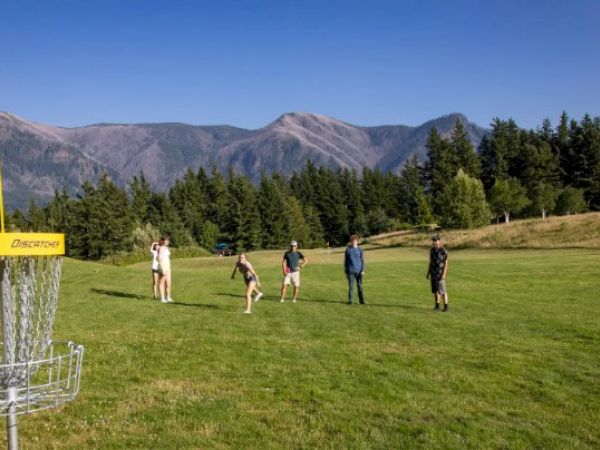 A group of people is playing disc golf on a grassy field with a disc golf basket nearby and mountains in the background.