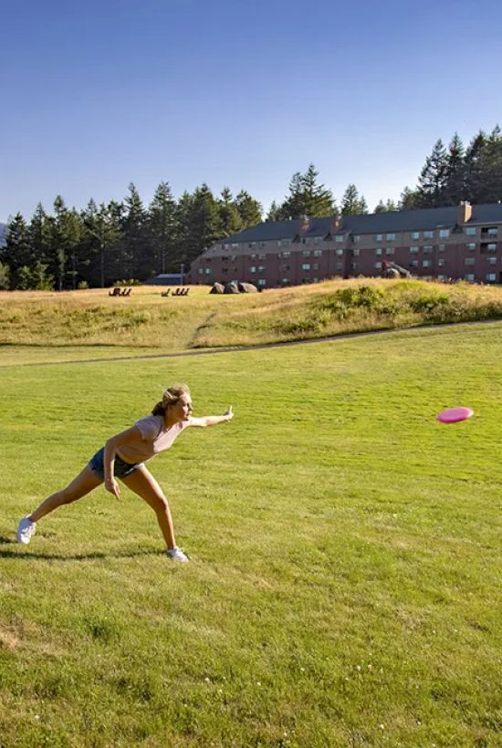 A person is playing with a frisbee on a lush green lawn near a large building, surrounded by trees under a clear blue sky.
