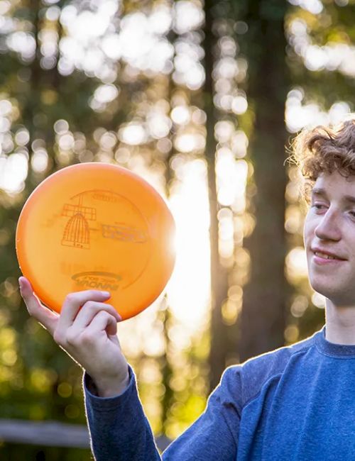 A person holds an orange Frisbee in a wooded area, enjoying the sunny day. The sunlight filters through the trees, creating a warm glow.