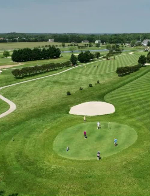 The image shows an aerial view of a golf course with people playing, lush green fairways, sand traps, and surrounding landscapes.