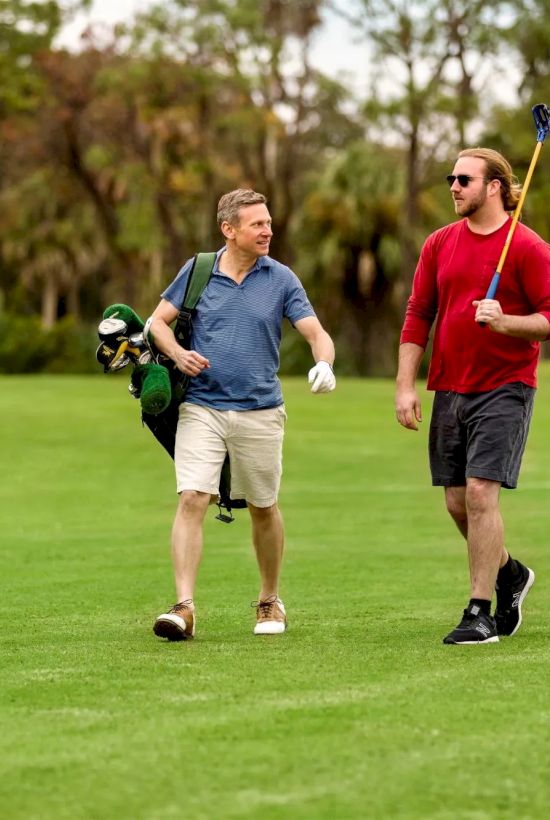 Two people walking on a golf course; one carries clubs, the other, a rake or similar pole. They are engaged in conversation.