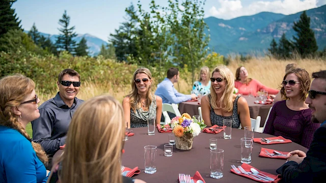 A group of people sitting at a round table outdoors, with a brown tablecloth, red napkins, and a mountain view in the background.